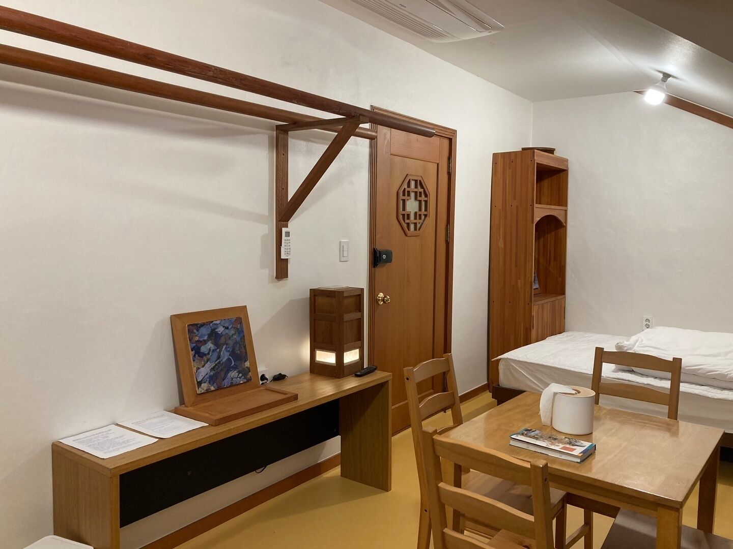 A compact room with a bed, a desk, and a chair. There's a wooden door with a decorative panel, a small shelf, and a wall-mounted light fixture.