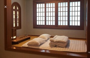 Una cama bien hecha con almohadas y un edredón acolchado, un pequeño estante con objetos, y un ventanal con diseño de cuadrícula.