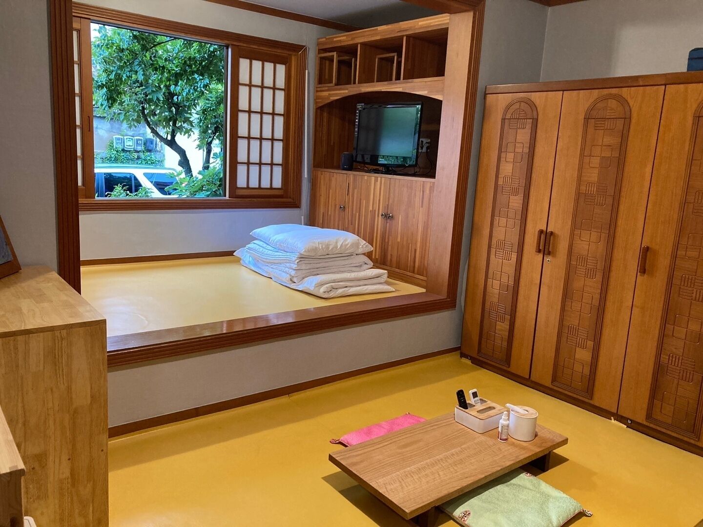 A room with a tatami mat floor, a low wooden bed with a window, a television on a shelf, and wooden cabinets.
