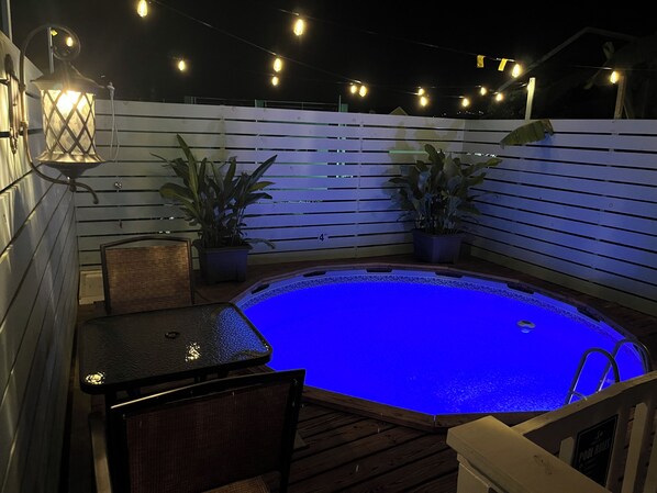 Un jacuzzi iluminado con luz azul, rodeado por una terraza de madera y plantas en macetas.