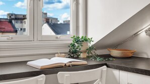 Une petite cuisine avec une fenêtre, des chaises blanches, une plante et un bol posé sur le comptoir.