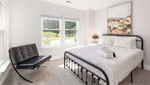 A bedroom with a bed, a black leather chair, a window with blinds, and a painting on the wall.