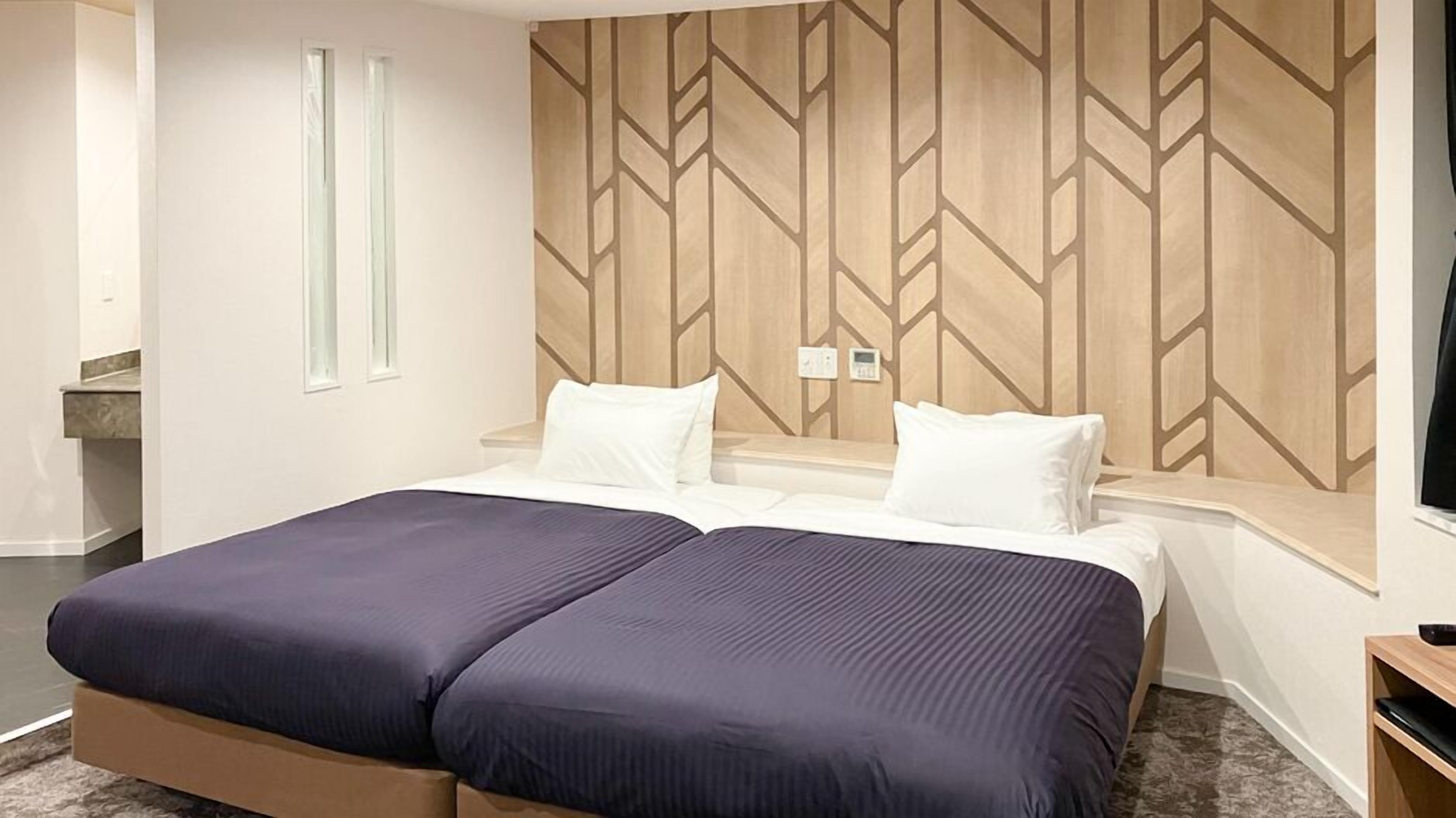 Une chambre d’hôtel comprenant un lit, deux tables de chevet, un thermostat fixé au mur et une fenêtre.