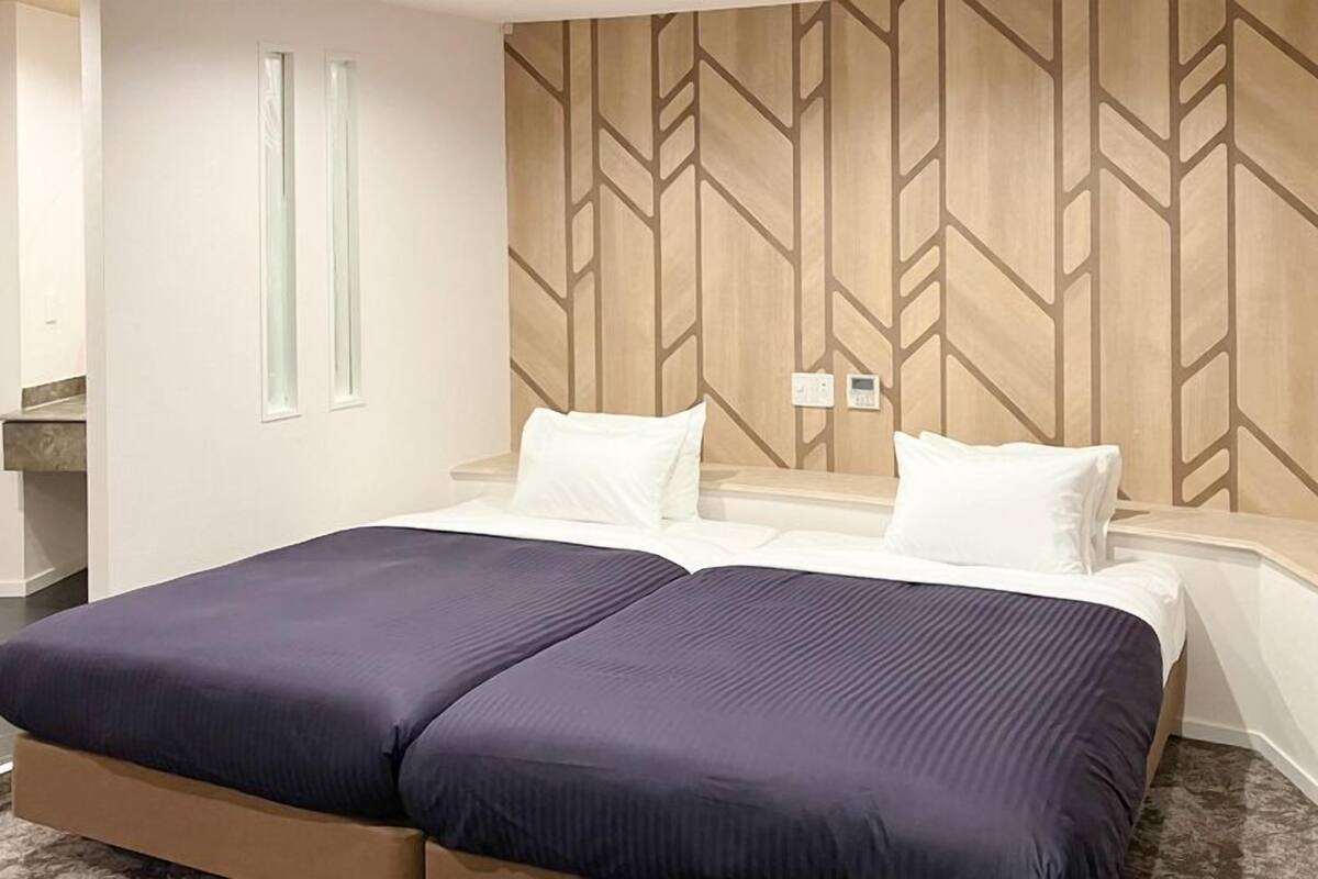 Une chambre d’hôtel comprenant un lit, deux tables de chevet, un thermostat fixé au mur et une fenêtre.