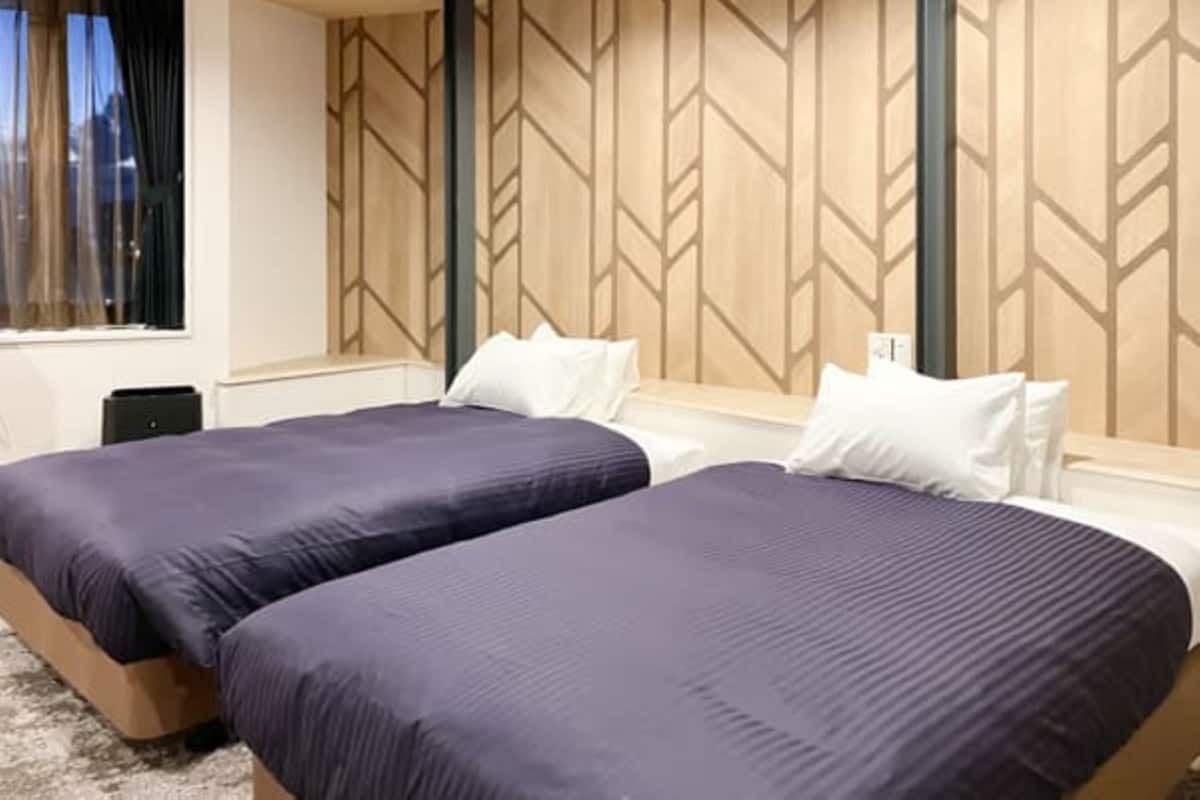 Une chambre d’hôtel avec deux lits, une fenêtre avec des rideaux, un panneau mural en bois et une petite table.