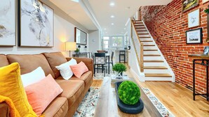 A spacious living area with a brown leather sofa, a wooden coffee table, and a staircase leading to an upper level. - Bright & Beautiful 4BR Home + Parking, Walk to Harbor & Stadiums (Baltimore)