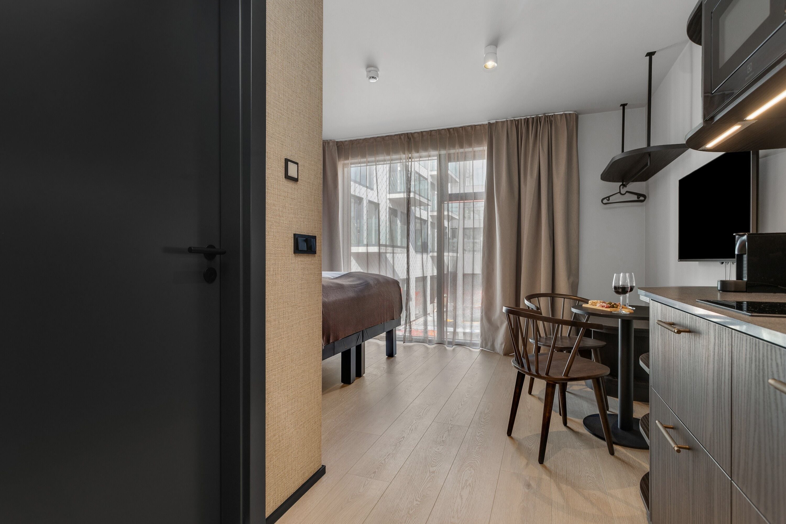 A modern hotel room with a bed, a dining table with chairs, a kitchen area, and a large window with curtains.