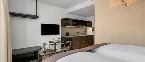 A modern hotel room with a bed, a small dining table, a TV, and a kitchenette.