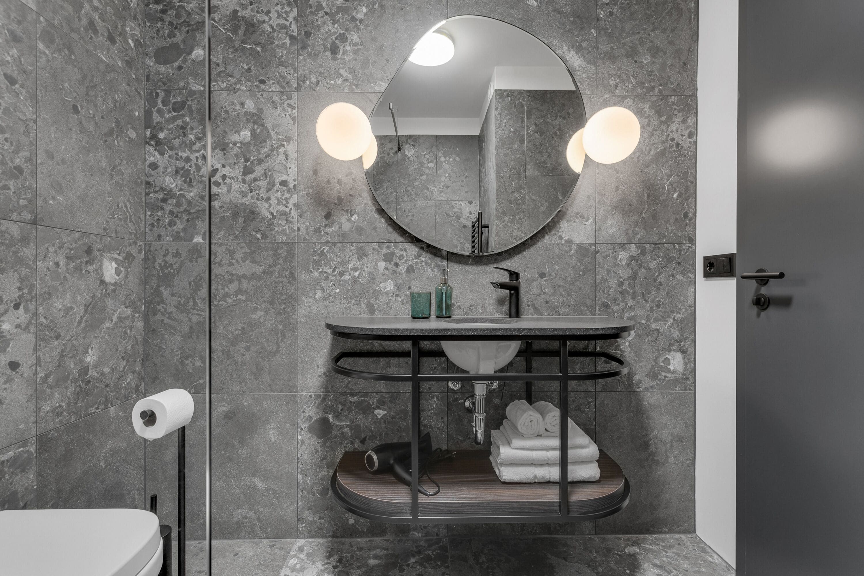Un baño con pared de mármol, un espejo redondo con luces, un lavabo con toallero y un inodoro.