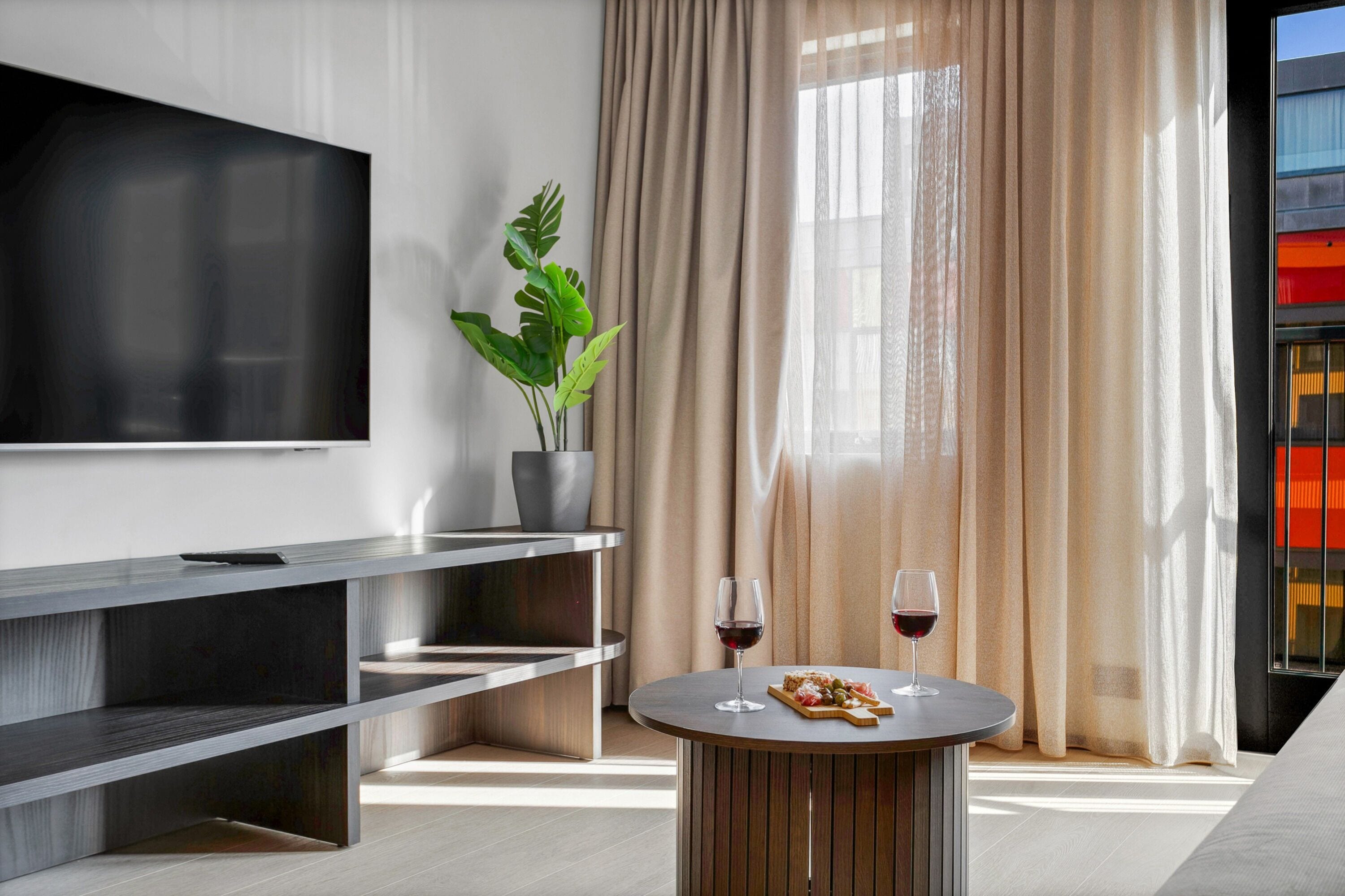 A modern living room with a flat-screen TV, a round wooden coffee table with a glass of red wine and a plate of food, a potted plant, and a large window with beige curtains.