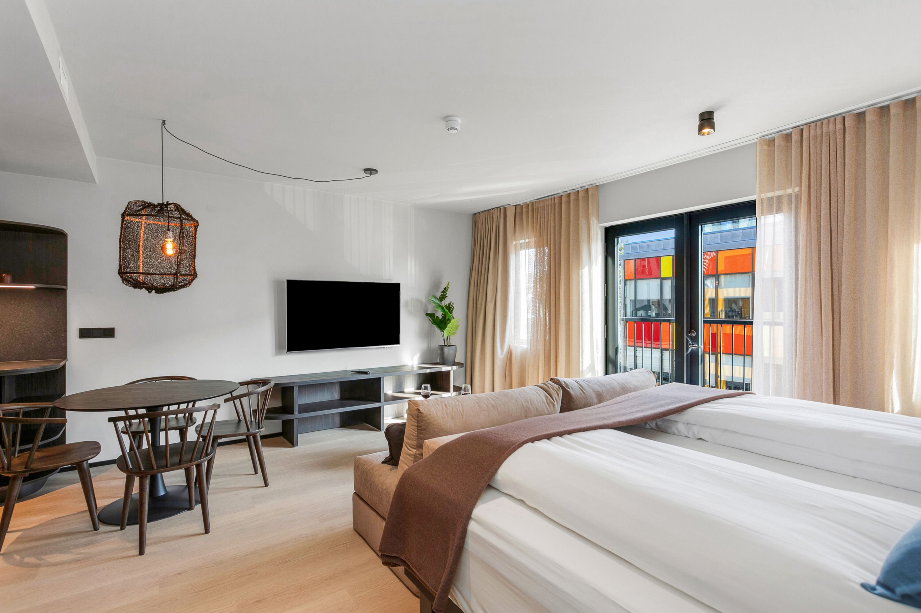 Habitación de hotel moderna con una cama grande, televisor, zona de comedor con sillas y vista a un edificio colorido por la ventana.