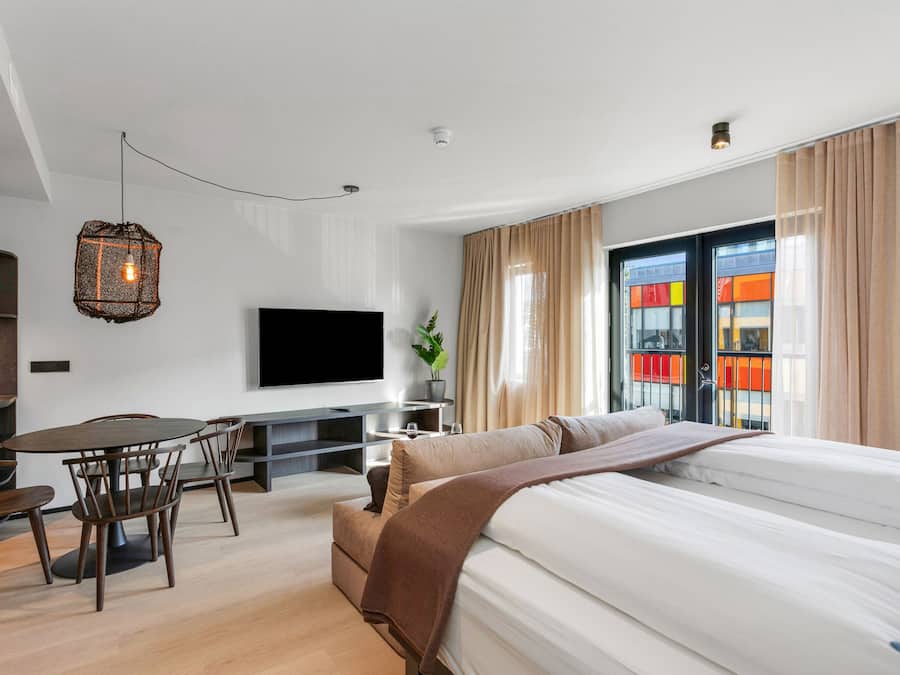 Habitación de hotel moderna con una cama grande, televisor, zona de comedor con sillas y vista a un edificio colorido por la ventana.