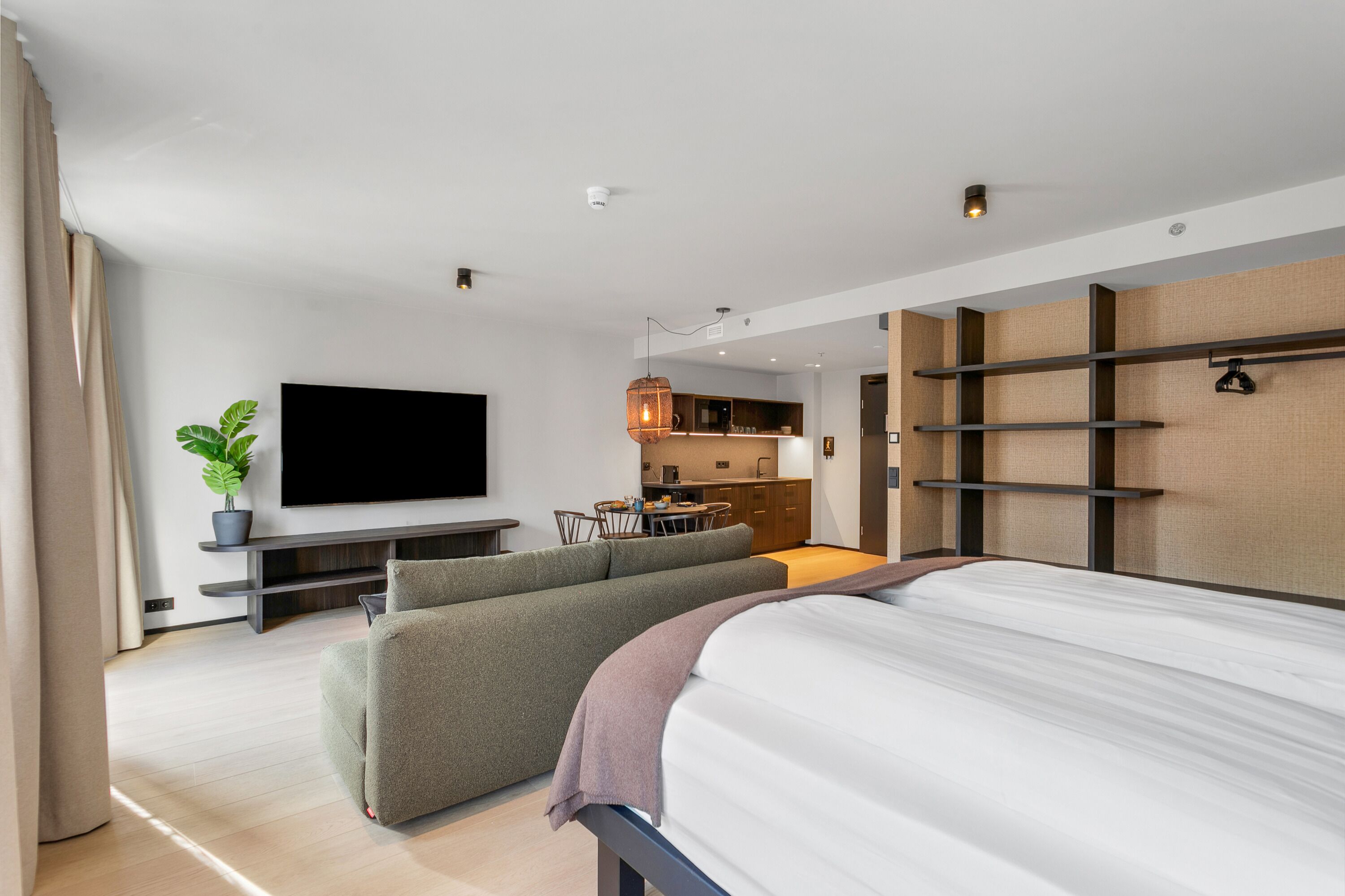 A modern hotel room with a large bed, a sofa, a flat-screen TV, a dining area, and a kitchen.
