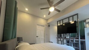 A hotel room with a bed, a ceiling fan, a TV, and a kitchenette area. - Casita Heliconia- Jr. Suite#3 (Roseau)