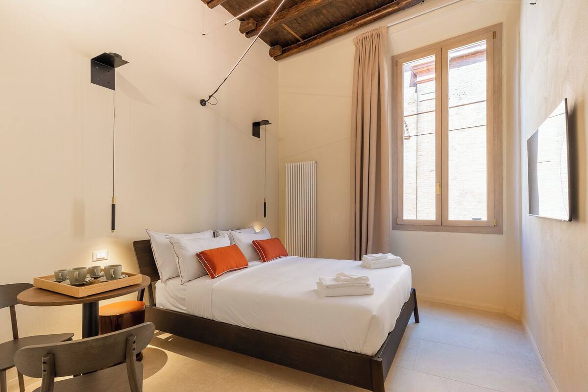 A bedroom with a bed, a bedside table with a tray of cups, a chair, a radiator, and a window with curtains.