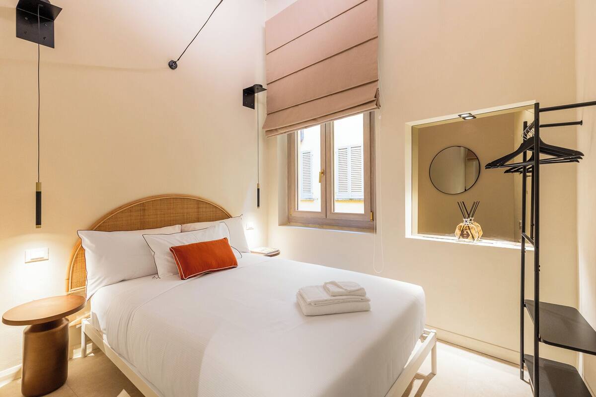 A bedroom with a bed, bedside table, window with blinds, and a wall-mounted mirror.