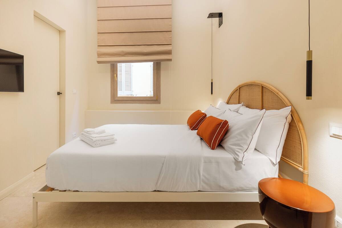 A neatly made bed with white linens and orange pillows, a wooden headboard, and a small window with blinds.