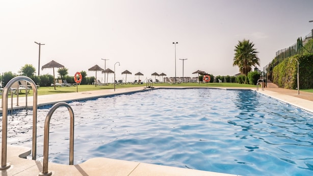 A swimming pool with lounge chairs and umbrellas, surrounded by greenery and a clear sky. - Modern Apartment with Sea & Mountain Views – Small Oasis, Manilva (Manilva)