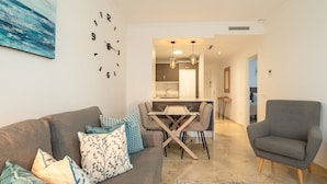 A modern living room with a grey sofa, a dining table, and a grey armchair. A kitchen is visible in the background with pendant lighting. - Modern Apartment with Sea & Mountain Views – Small Oasis, Manilva (Manilva)