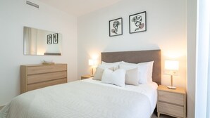 A hotel room with a large bed, a wooden dresser, a nightstand with a lamp, and two framed artworks on the wall. - Modern Apartment with Sea & Mountain Views – Small Oasis, Manilva (Manilva)
