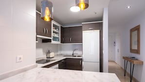 A modern kitchen with marble countertops, a built-in oven, and a refrigerator. There are wooden cabinets and a small dining area with a table and chairs. - Modern Apartment with Sea & Mountain Views – Small Oasis, Manilva (Manilva)