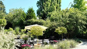 An outdoor seating area with tables and chairs under a canopy, surrounded by various plants and trees.