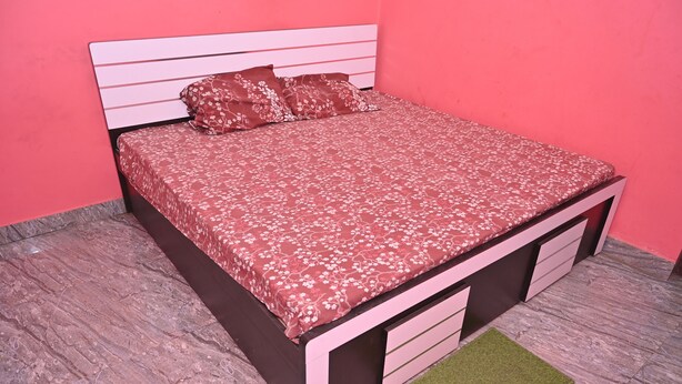 A bed with a floral bedspread and matching pillows, a headboard, a footboard, and a small green rug on a tiled floor. - The Ganga Guest House (Varanasi)