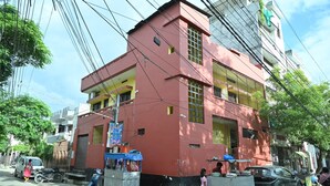 A multi-story building with a red facade, a gated entrance, and a small shop on the ground floor. - The Ganga Guest House (Varanasi)