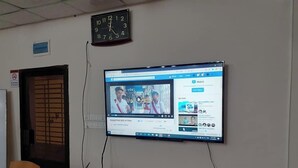 A room with a desk, a computer monitor displaying a video, a clock, and a door with a barred window. - Haor Bilash Guest House (Sunamganj)