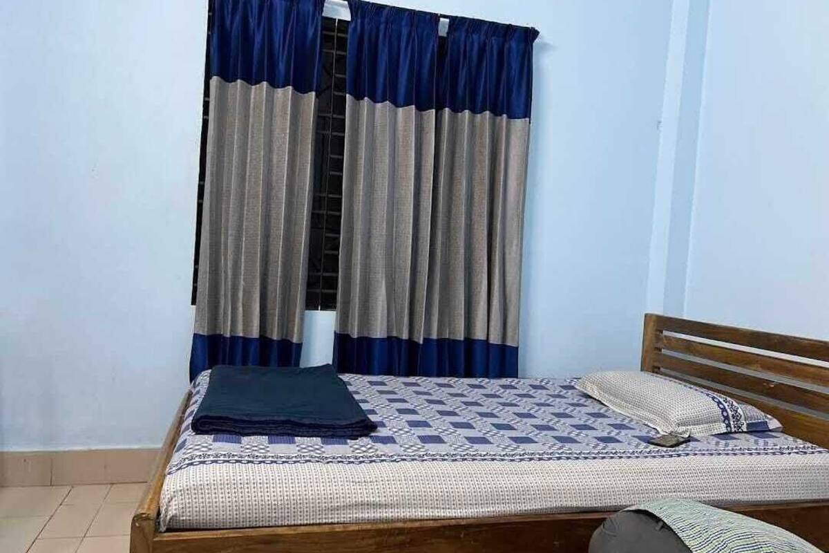 Ein Einzelbett mit einem gemusterten Bettüberwurf, einem Holz-Kopfteil und einem Fenster mit blau-grauen Vorhängen.