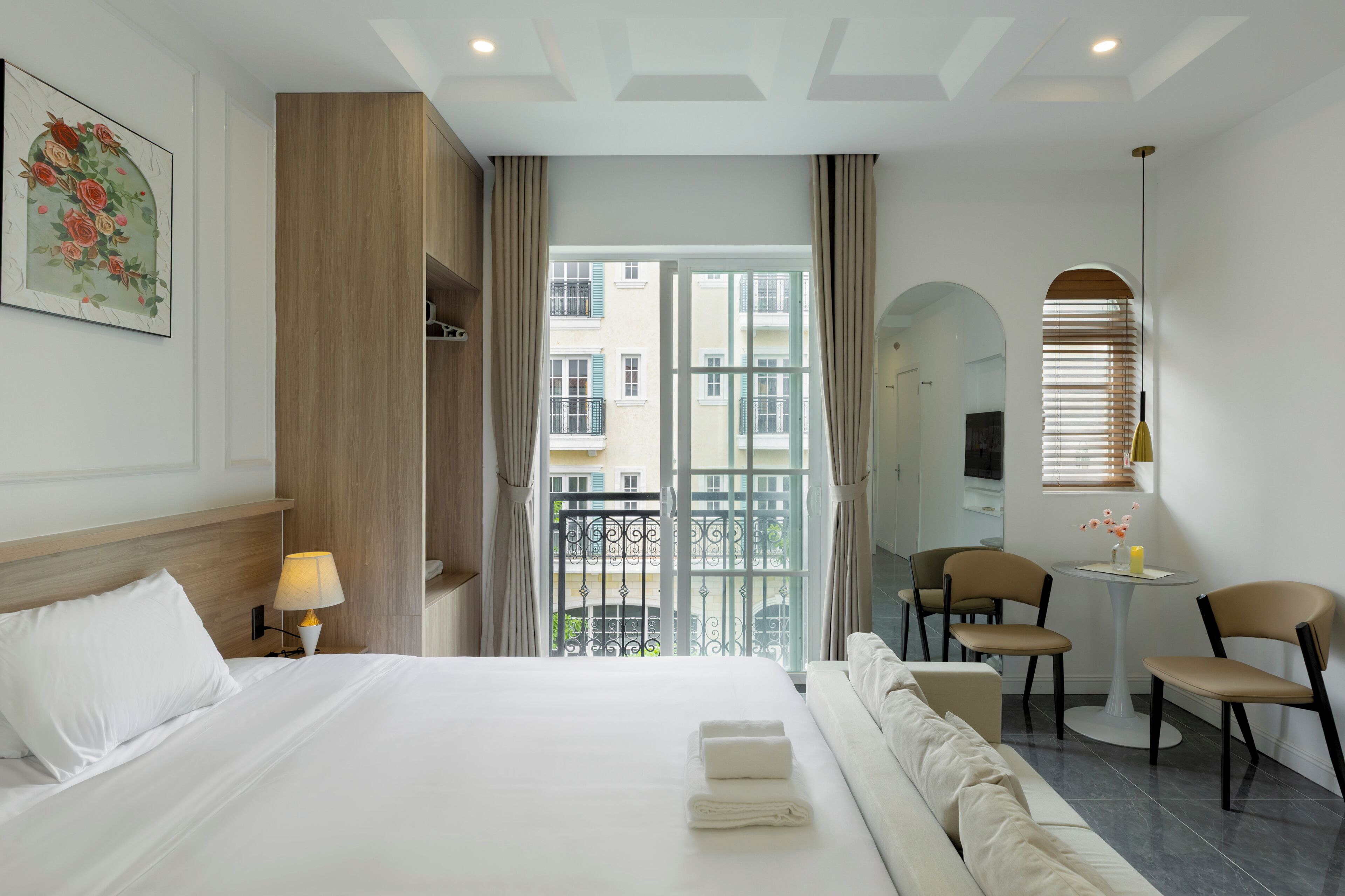 A hotel room with a bed, a desk, two chairs, a small table with a vase, and a balcony view.