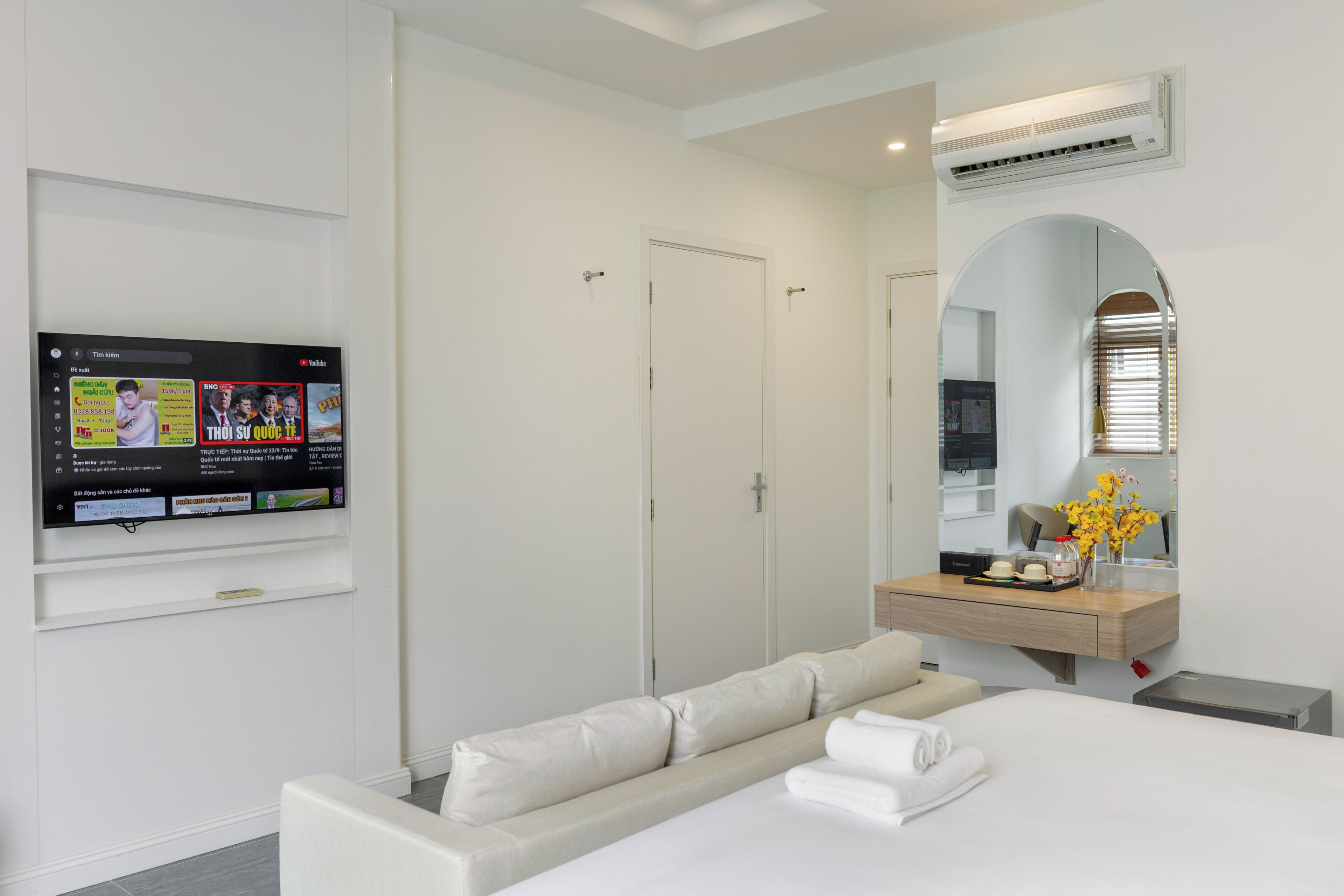 A hotel room with a bed, a sofa, a TV mounted on the wall, a mirror, and a small table with a vase of flowers.