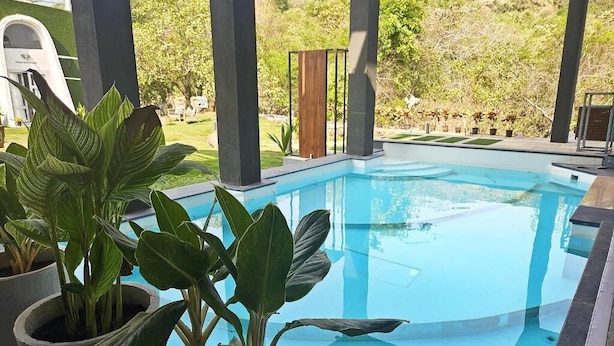 A swimming pool with potted plants in the foreground, surrounded by a covered area with pillars and a view of a landscaped garden. - Nyast Valley Resort (Arpora)