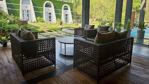 A covered outdoor seating area with wicker furniture and a view of a green building with arched windows. - Nyast Valley Resort (Arpora)