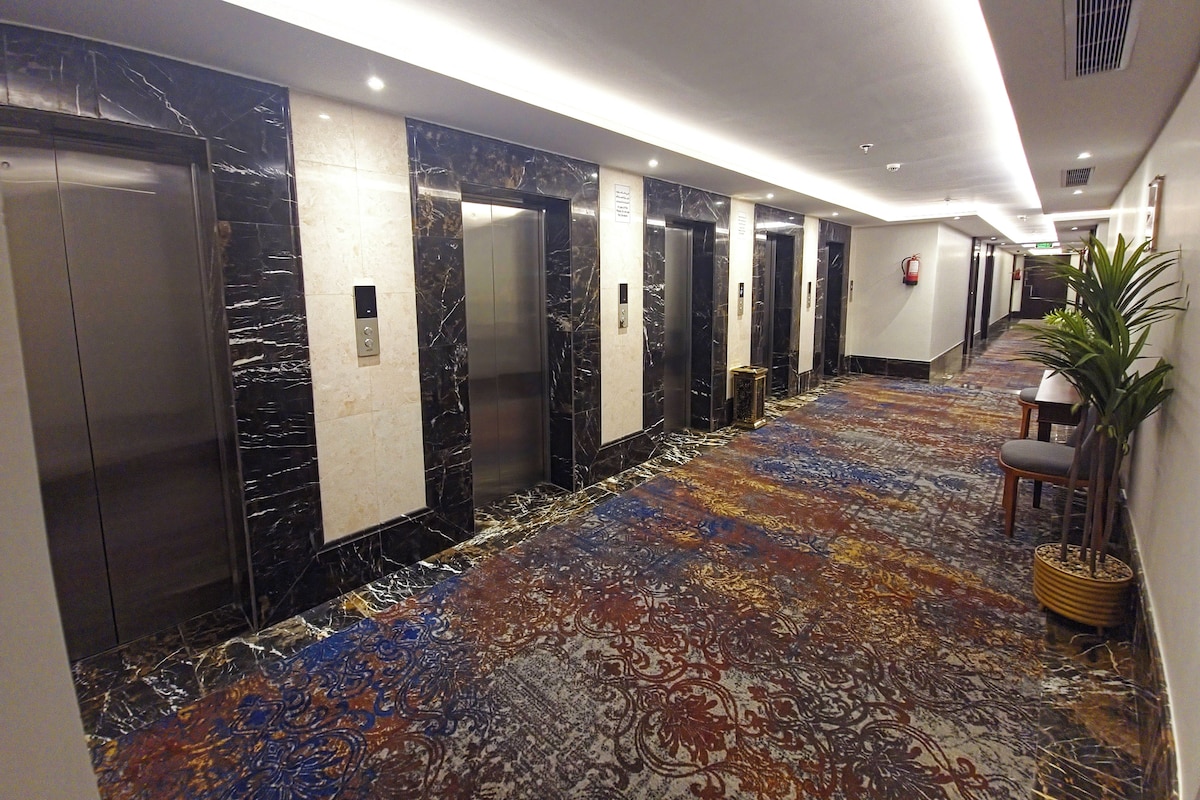 A hotel corridor with elevators, a patterned carpet, and a potted plant.