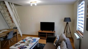 A living room with a TV, a sofa, a coffee table with magazines, a lamp, and a window with blinds. - Cosy gîte in renovated farmhouse. Spacious bedroom + living room. Pets allowed (Weitbruch)