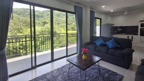 A modern living room with a large sliding glass door leading to a balcony, a dark sofa with blue cushions, a small wooden coffee table with a vase of flowers, and a kitchen area with a black backsplash and white cabinets. - Tropical Private Pool Villa Phuket (ตำบล ฉลอง)