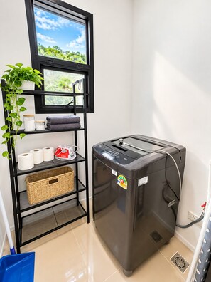 A laundry room with a black washing machine, a shelf with towels, a basket, and a window with a view of greenery. - Tropical Private Pool Villa Phuket (ตำบล ฉลอง)