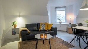 A modern loft apartment with a black leather sofa, a round coffee table, and a dining table with chairs. There is a window with a view of buildings outside, a radiator, and a potted plant on a shelf. - Scandinavian Charm in Aalborg (Aalborg)