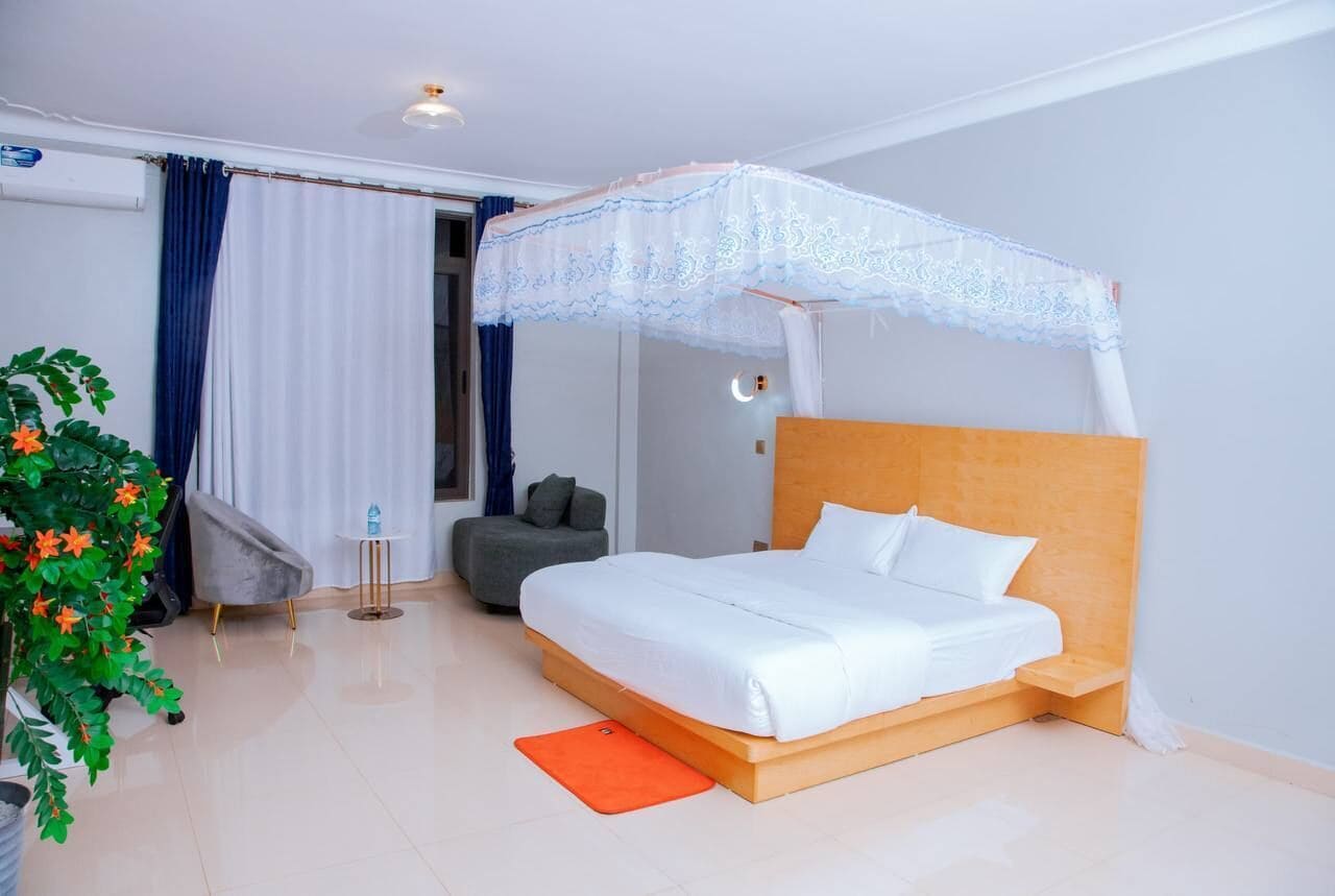 Un dormitorio moderno con una cama con dosel, un sillón gris, una mesita pequeña y una planta en maceta.