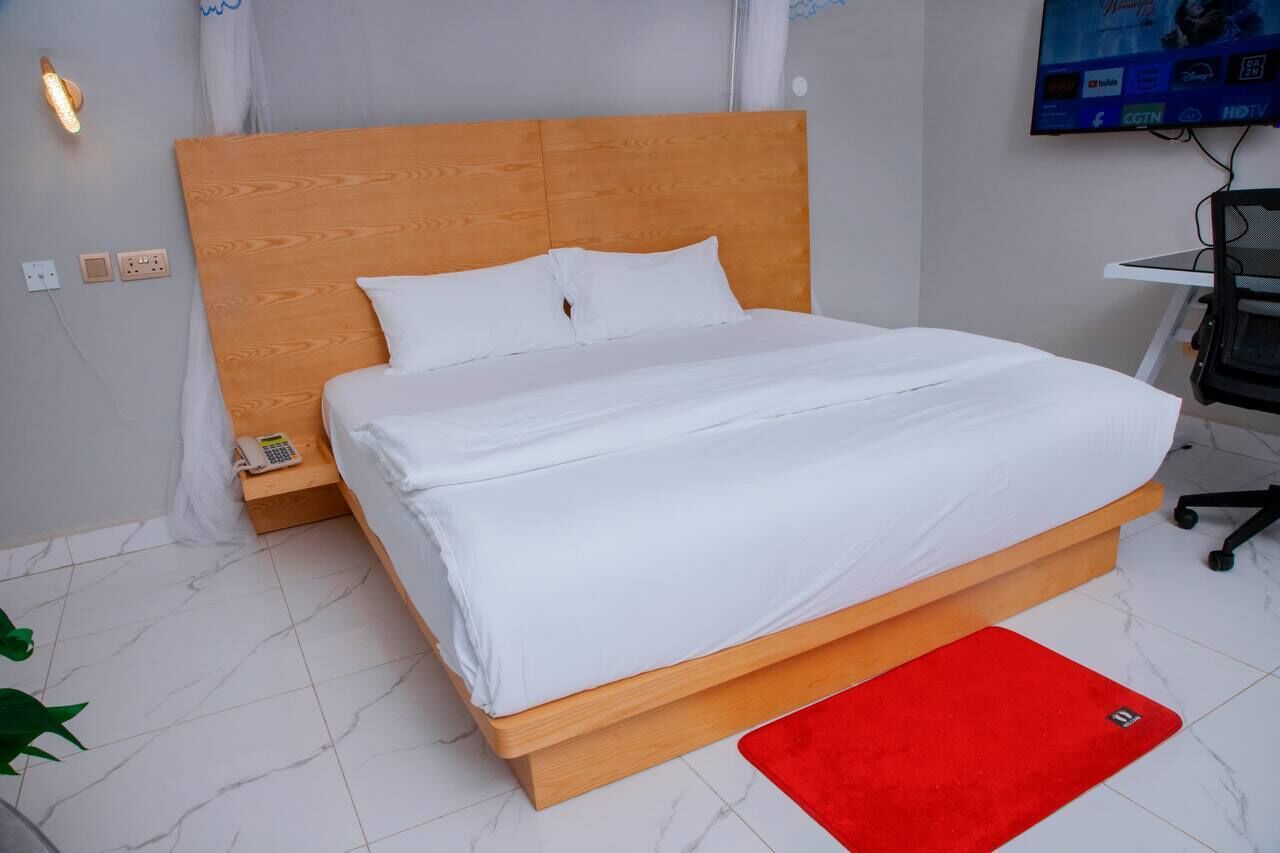 Una cama bien tendida con ropa blanca, cabecera de madera y un tapete rojo sobre suelo de baldosas.