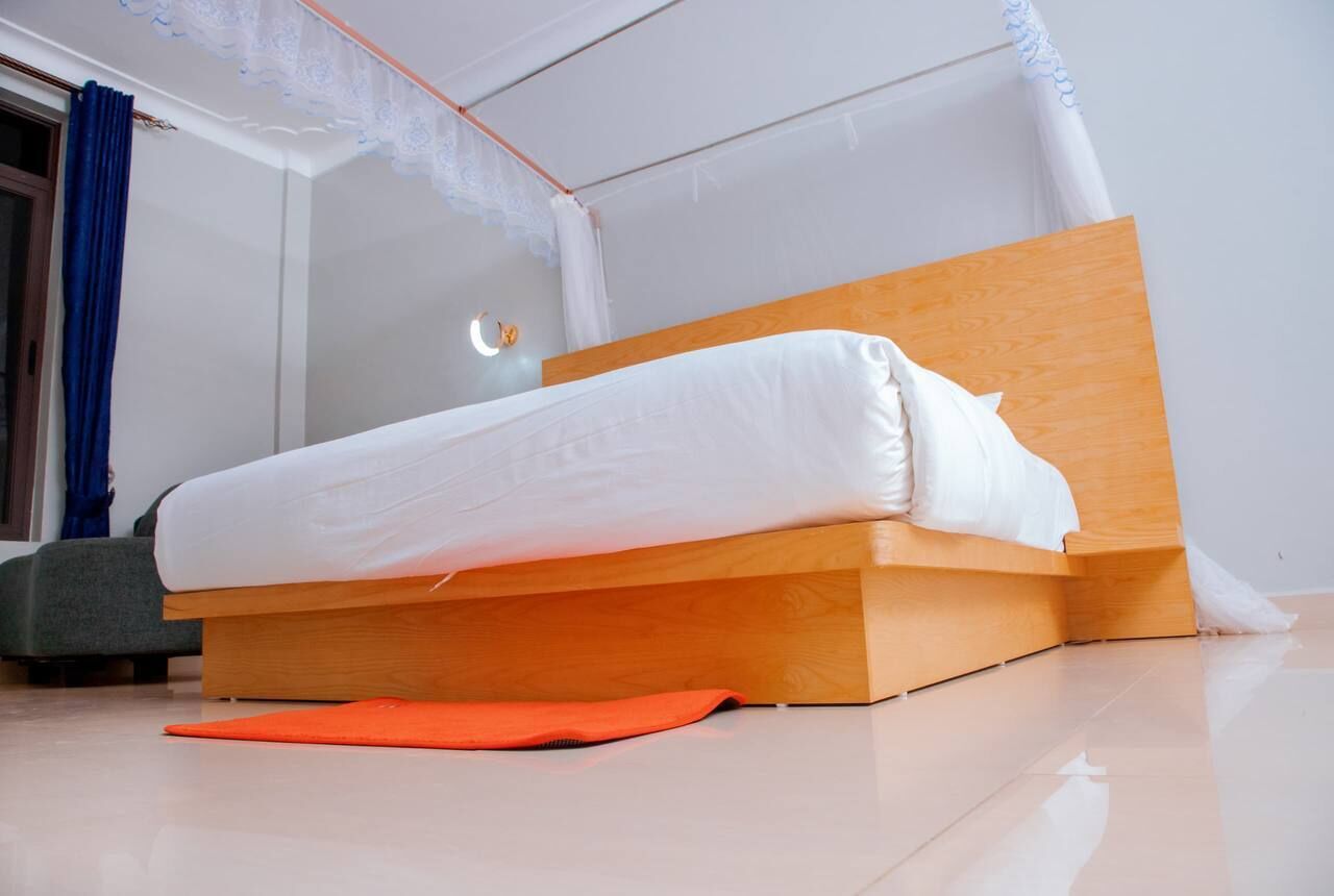 Uma cama de solteiro com cabeceira de madeira e roupa de cama branca num quarto com soalho em mosaico e janela com cortinas azuis.