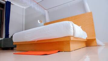 Uma cama de solteiro com cabeceira de madeira e roupa de cama branca num quarto com soalho em mosaico e janela com cortinas azuis.