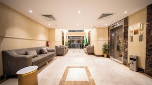 Egy szállodai előtér, amelyben recepció, kanapék, szemeteskosár és lift található.