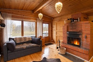 A cozy cabin interior with a fireplace, wooden walls, a sofa, and a hanging light fixture. - Aurora River Cabin Ivalo – Hot Tub, Sauna & Cold Plunge (Inari)