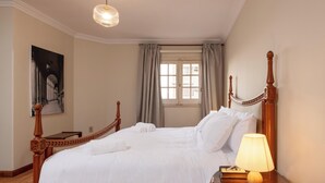 A neatly made bed with white linens, a wooden headboard, a bedside table with a lamp, and a painting on the wall. - Kennah Lascaic | 3BR Near Korba & Metro, Heliopolis (Cairo)