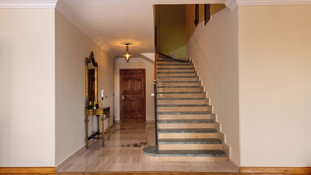A hallway with a wooden floor, a wooden door, and a staircase with a handrail. - Kennah Lascaic | 3BR Near Korba & Metro, Heliopolis (Cairo)