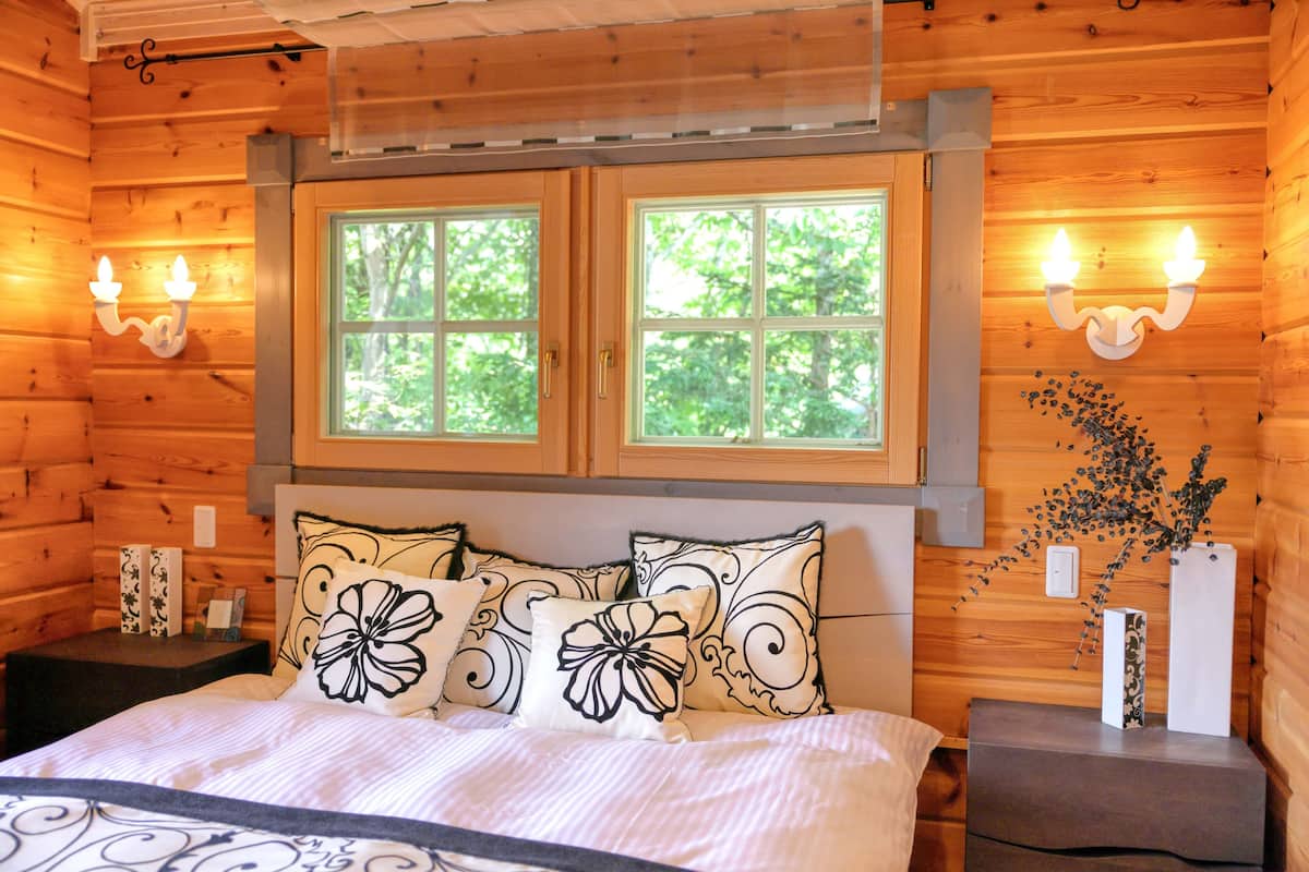 A wooden cabin bedroom with a bed, two windows, and wall-mounted lights.