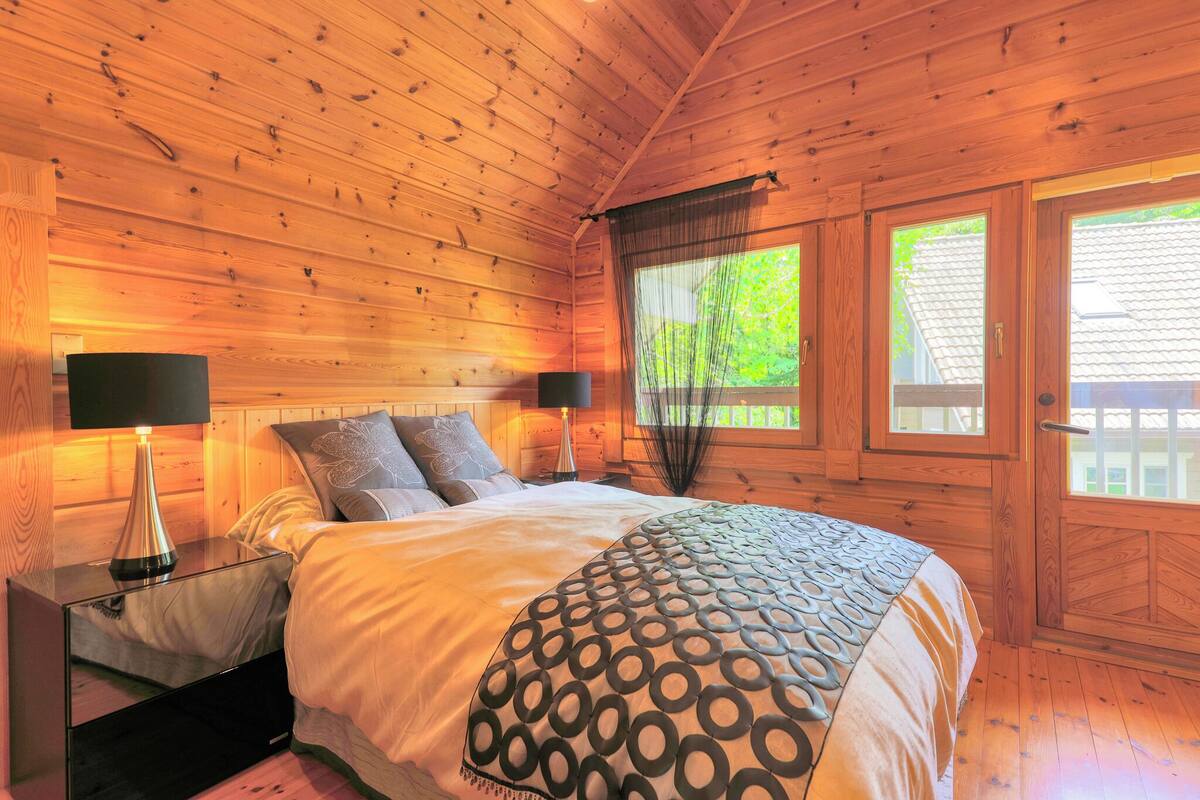 A wooden cabin bedroom with a bed, two bedside tables with lamps, a window with curtains, and a door.