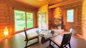 A wooden cabin interior with a glass table, two chairs, a bench, and a large window offering a view of the outdoors.